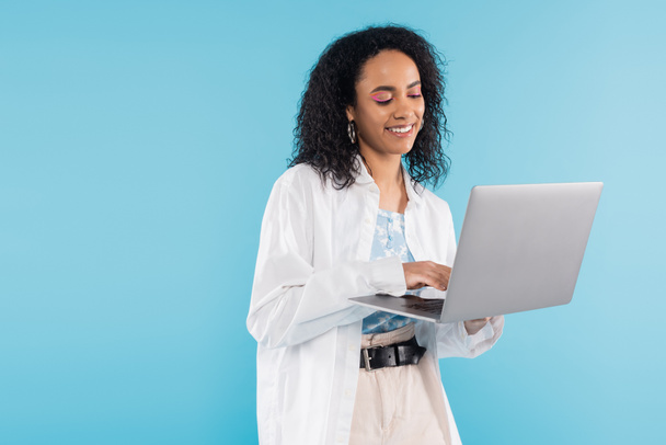 brunette african american woman in white shirt smiling while using laptop isolated on blue - Zdjęcie, obraz