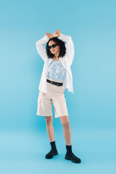 full length of young african american woman in shorts and sunglasses posing with hands above head on blue background - 写真・画像