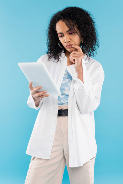 вдумчивая африканская американка в белой стильной рубашке смотрит на цифровой планшет, изолированный на синий - Фото, изображение