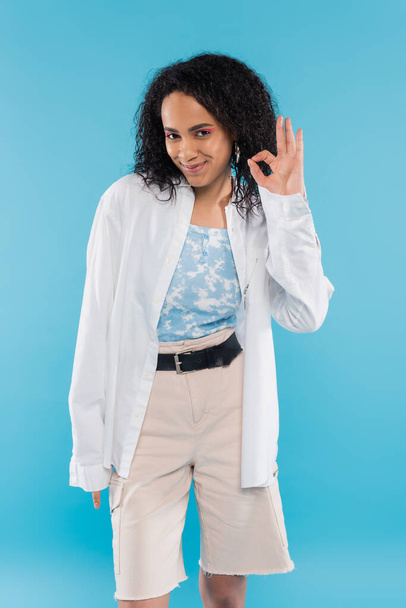 pleased african american woman in trendy clothes looking at camera and showing okay sign isolated on blue - Photo, Image