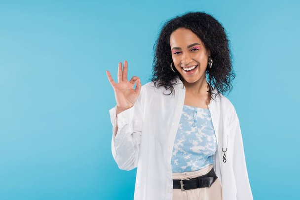 overjoyed african american woman in stylish shirt smiling at camera and showing okay sign isolated on blue - Фото, изображение