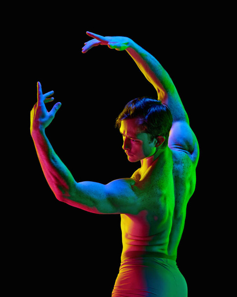 Ruumis hehkuu. Nykytaiteen baletti. Nuori joustava urheilullinen mies, näyttelijä poseeraa kädet tumman studion taustalla neonvalolla. Moderni balettitanssija. Grace, taide, ammattiurheilun käsite - Valokuva, kuva