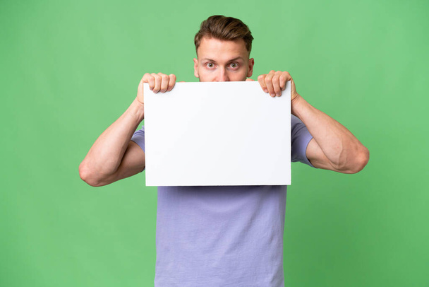 Young blonde caucasian man over isolated background holding an empty placard and hiding behind it - Фото, зображення