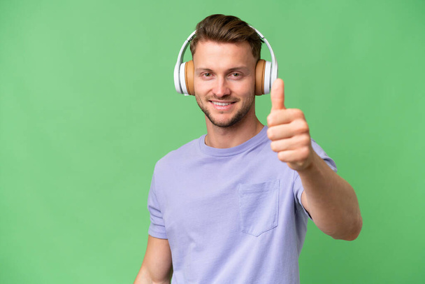 jong blond Kaukasische man over geïsoleerde achtergrond luisteren muziek en met duim omhoog - Foto, afbeelding
