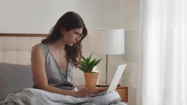 Молода брюнетка сидить на ліжку з ноутбуком на колінах і швидко друкує щось. Молода жінка здивована ноутбуком, який замерзає під час роботи. Високоякісні FullHD кадри
 - Кадри, відео
