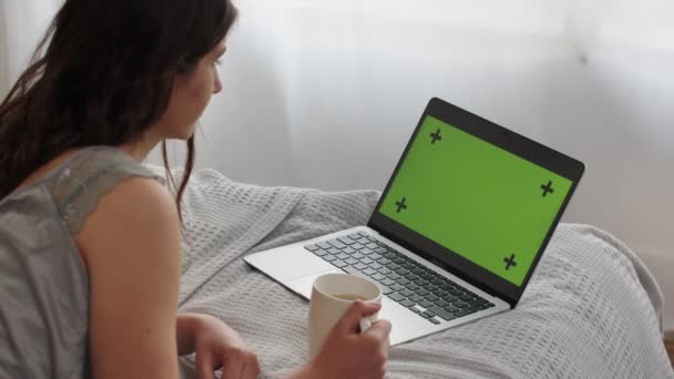 Długowłosy brunetka leży na łóżku z filiżanką herbaty i patrzy na coś na jej laptopie. Kobieta leży przed laptopem z zielonym ekranem i okresowo naciska klawisze. Wysoka jakość - Materiał filmowy, wideo