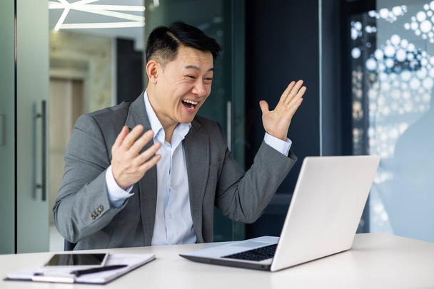 Successful asian investor financier celebrating victory and triumph, mature boss in business suit working inside office with laptop, man received online win news. - Photo, image