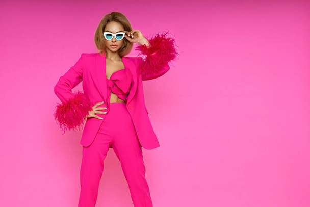 Modelo de alta moda. Mujer elegante está usando chaqueta de plumas de color rosa, pantalones y tacones altos sobre fondo rosa. Modelo femenino elegante posando en traje de lujo con estilo. - Foto, imagen
