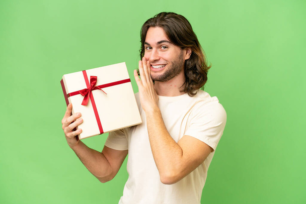 Junger gutaussehender Mann hält ein Geschenk über isoliertem Hintergrund und flüstert etwas - Foto, Bild