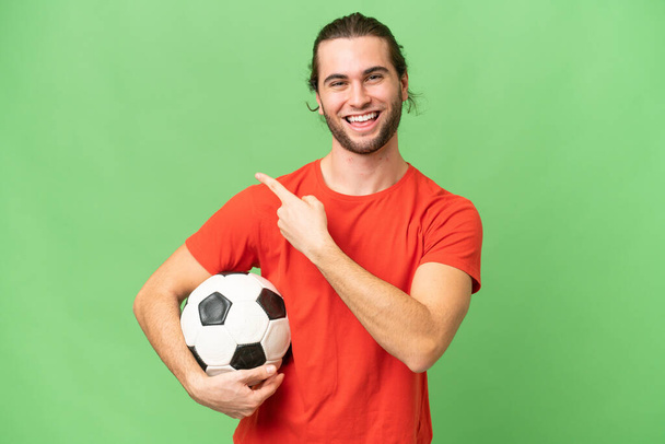 Mladý pohledný muž izolovaný na zeleném chroma pozadí s fotbalovým míčem a ukazuje na boční - Fotografie, Obrázek