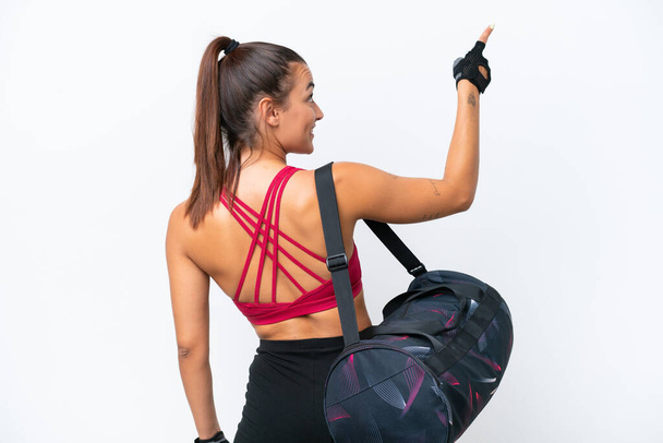 Jeune femme sportive avec sac de sport isolé sur fond blanc pointant vers l'arrière avec l'index - Photo, image