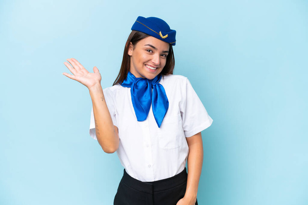 Avion hôtesse de l'air femme caucasienne isolée sur fond bleu saluant avec la main avec expression heureuse - Photo, image