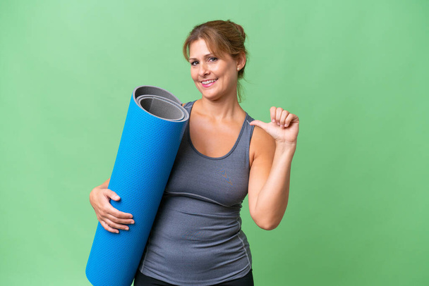 Middle-aged sport woman going to yoga classes while holding a mat over isolated background proud and self-satisfied - Foto, imagen