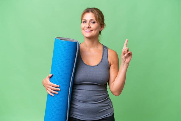 Middelbare leeftijd sport vrouw die naar yoga klassen terwijl het houden van een mat over geïsoleerde achtergrond tonen en tillen van een vinger in teken van de beste - Foto, afbeelding