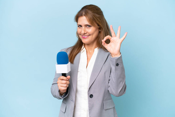 Middle-aged TV presenter woman over isolated blue background showing ok sign with fingers - Photo, Image