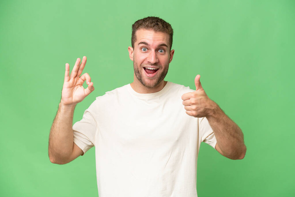 Jonge knappe blanke man geïsoleerd op groene chroma achtergrond met ok teken en duim omhoog gebaar - Foto, afbeelding
