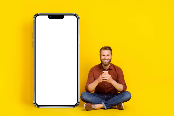 Full length photo of handsome guy sit floor use telephone empty space big electronic panel isolated on yellow color background. - Foto, imagen