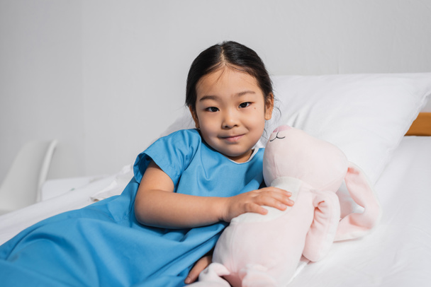 heureux asiatique fille embrassant jouet lapin et regarder caméra sur lit dans hôpital salle - Photo, image
