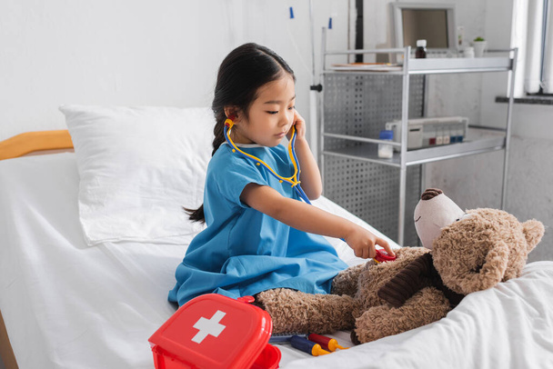 Küçük Asyalı kız hastane yatağında oyuncak steteskopla oyuncak ayıyı muayene ediyor. - Fotoğraf, Görsel