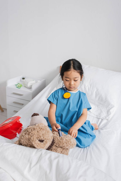 asiático niño en hospital bata haciendo inyección a teddy oso con juguete jeringa mientras jugando en cama en clínica - Foto, imagen