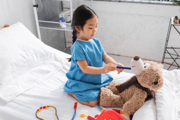 aasialainen tyttö sairaalan puku tilalla lelu ruisku lähellä nallekarhu pelatessaan sängyssä klinikalla - Valokuva, kuva