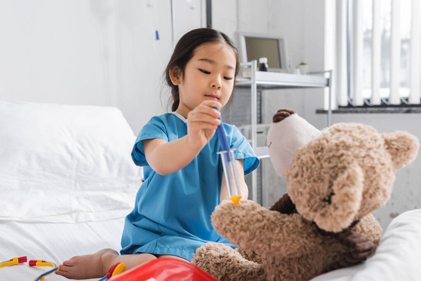 Hastane önlüklü küçük Asyalı çocuk oyuncak şırınga ile oyuncak ayıya iğne yapıyor.  - Fotoğraf, Görsel