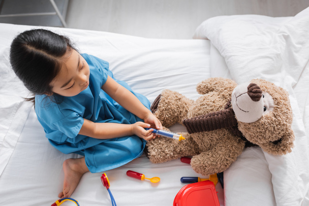 pohled shora na asijské dítě hrající si na nemocniční posteli a provádění injekce plyšovému medvídkovi s injekční stříkačkou - Fotografie, Obrázek