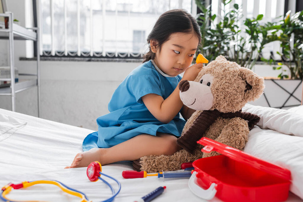 Ασιάτης με νοσοκομειακή ρόμπα εξετάζει αρκουδάκι με ωτοσκόπιο στο κρεβάτι σε παιδιατρική κλινική - Φωτογραφία, εικόνα