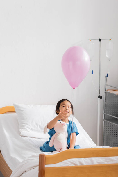 ásia menina sentado com brinquedo coelho no hospital cama e olhando para balão - Foto, Imagem