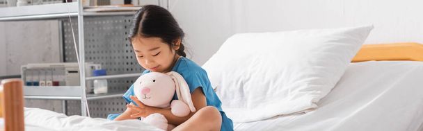 χαμογελαστό ασιατικό κορίτσι αγκαλιάζει παιχνίδι λαγουδάκι, ενώ κάθεται στο κρεβάτι στο θάλαμο του νοσοκομείου, πανό - Φωτογραφία, εικόνα