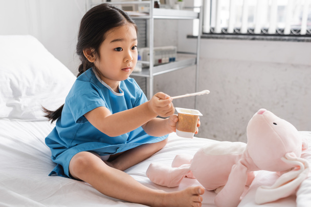 blootsvoets aziatisch meisje houden heerlijke yoghurt in de buurt speelgoed bunny terwijl zitten op ziekenhuis bed - Foto, afbeelding