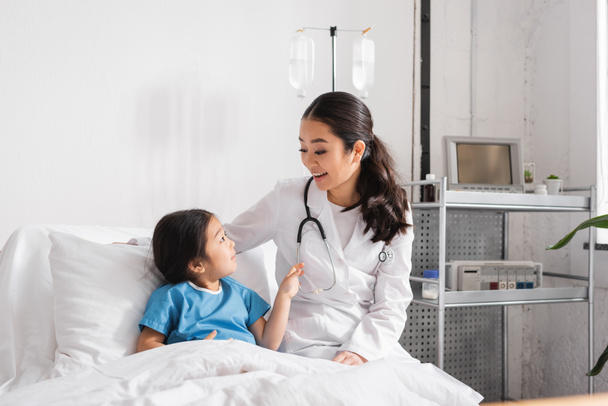 happy asian doctor with stethoscope talking to girl on bed in hospital ward - Foto, imagen