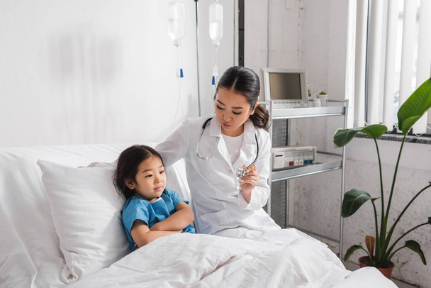 young doctor showing stethoscope to asian girl sitting on hospital bed with folded arms - Foto, imagen