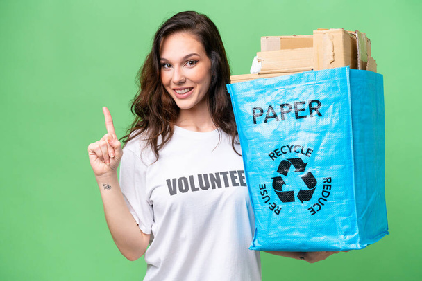 指を持ち上げながら解決策を実現しようとする隔離された背景をリサイクルする紙でいっぱいのリサイクルバッグを持っている若い白人女性 - 写真・画像