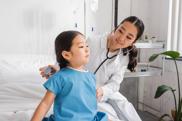 positiver Arzt mit Stethoskop untersucht asiatisches Kind im Krankenhauskittel sitzend auf Bett in Klinik - Foto, Bild