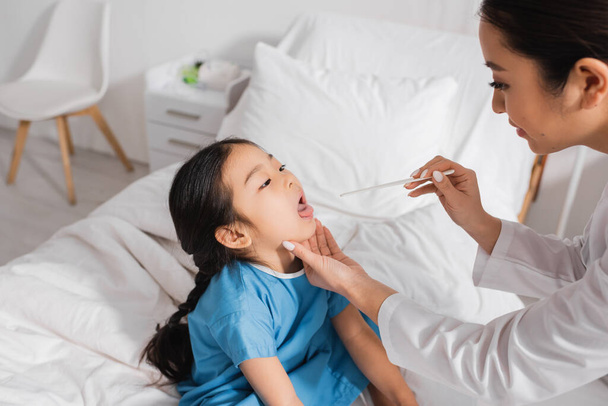 asian girl sitting with open mouth near pediatrician with tongue depressor in hospital ward - Фото, зображення