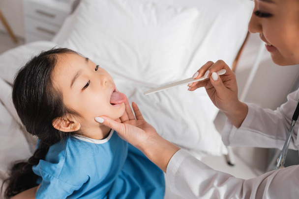 asian girl opening mouth near smiling doctor with tongue depressor in hospital ward - Photo, Image