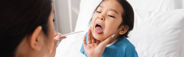 niño asiático con la boca abierta cerca del médico con depresor de la lengua en primer plano borroso en la sala del hospital, bandera - Foto, Imagen