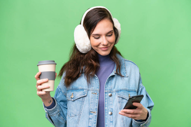 Young caucasian woman wearing winter muffs over isolated background holding coffee to take away and a mobile - Foto, Imagem