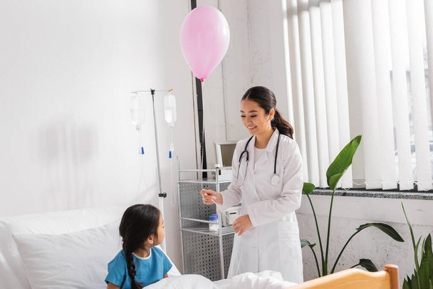 cheerful asian pediatrician holding festive balloon near little patient sitting on bed in clinic - Fotó, kép