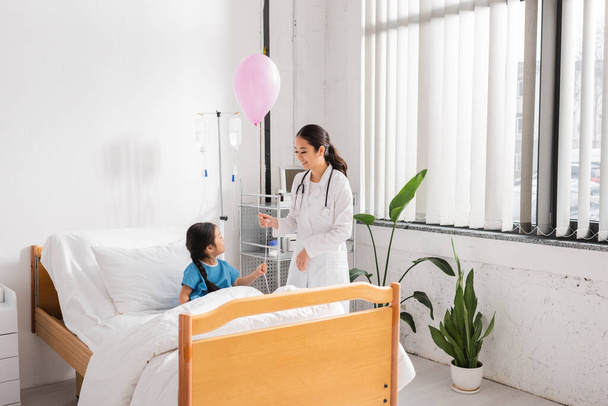 sonriente asiático doctor holding festivo globo cerca chica sentado en cama en moderno hospital sala - Foto, imagen