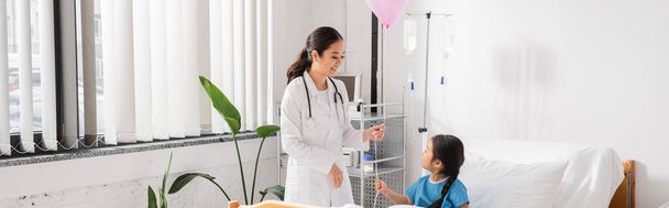 happy asian doctor in white coat holding festive balloon near kid in modern pediatric clinic, banner - Photo, image