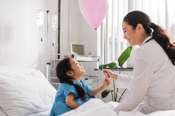 side view of young pediatrician giving festive balloon to overjoyed asian girl in hospital ward - Φωτογραφία, εικόνα