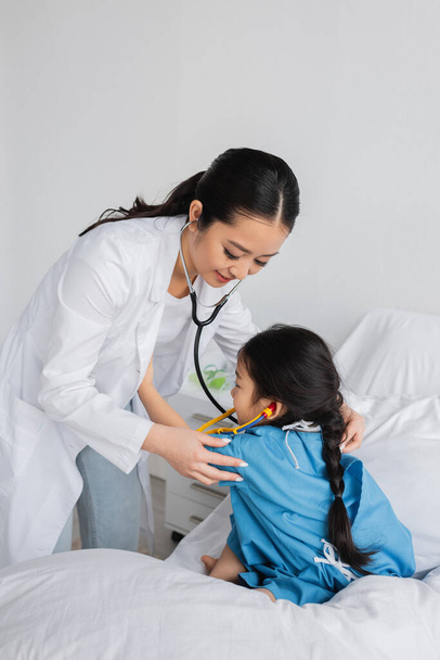 glimlachende aziatische kinderarts met stethoscoop onderzoeken kind in ziekenhuis toga zittend op bed in kliniek - Foto, afbeelding