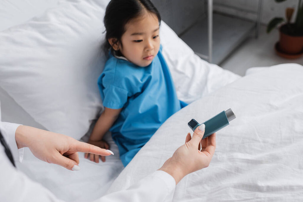 Doktor, hastane yatağında oturan gözü dönmüş Asyalı kızın yanında astım spreyini işaret ediyor. - Fotoğraf, Görsel