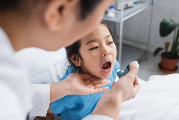 ziek aziatisch meisje opening mond in de buurt wazig kinderarts met inhalator in ziekenhuis afdeling - Foto, afbeelding