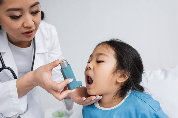 νεαρός Ασιάτης γιατρός κρατώντας εισπνευστήρα κοντά σε μικρό κορίτσι με ανοιχτό στόμα σε παιδιατρική κλινική - Φωτογραφία, εικόνα