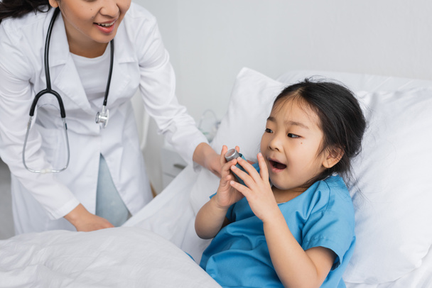 kleine asiatische Mädchen mit offenem Mund hält Inhalator in der Nähe Arzt in Kinderklinik - Foto, Bild