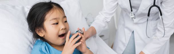 Ασιάτης παιδί κρατώντας εισπνευστήρα και το άνοιγμα του στόματος κοντά στο γιατρό σε λευκό παλτό στο θάλαμο του νοσοκομείου, πανό - Φωτογραφία, εικόνα