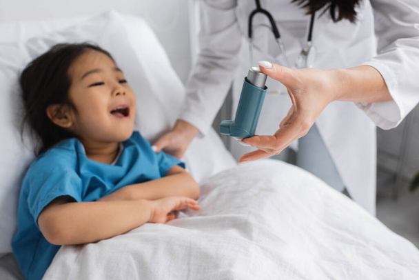 homályos gyermekorvos kezében inhalátor közel ázsiai gyermek nyitó száj kórházi ágyon - Fotó, kép